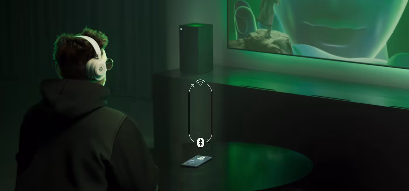 A man sitting on a couch with Arctis Nova 5 headphones on his ears; in front of him is a table with a phone on it. The man is looking at a distant TV, next to which stands a PS5 console.