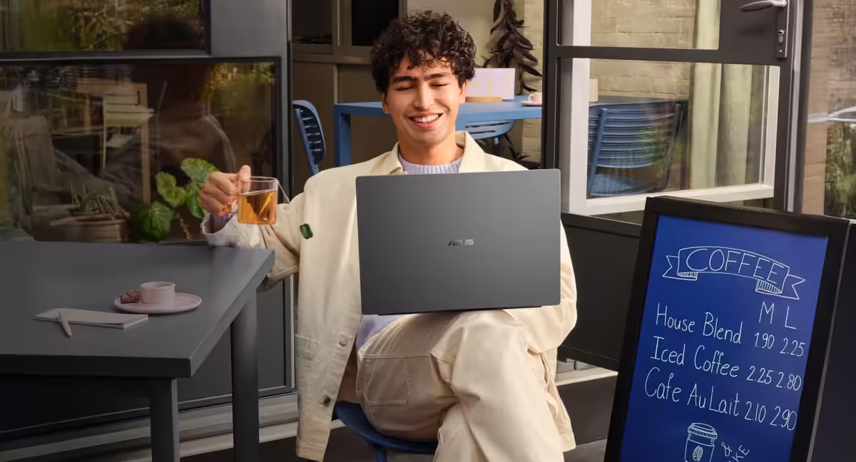 A man seated in a park, sipping coffee while working on a Matte Gray ASUS Vivobook S14.