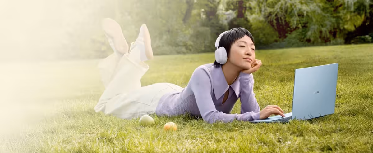 A woman lying on the grass using a light blue ASUS Vivobook S14 in the morning light.