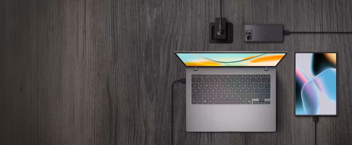 A top-down view of a clean, wooden workspace featuring a laptop, tablet, and smartphone connected by charging cables.