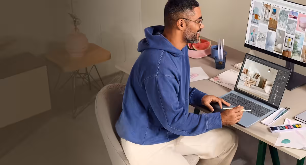 A person sitting at a desk using light blue ASUS Vivobook S14 connected to an external monitor, working on creative content with sketches, photos, and notes displayed on the wall.