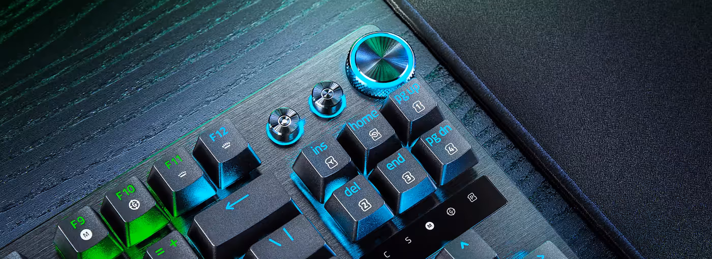Close-up of a mechanical keyboard with black keys, blue and green backlit labels, and a black mouse pad to the right.