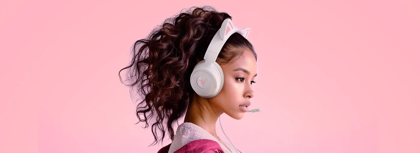 Young woman with curly hair wearing pink and white bunny-eared headphones with a microphone, against pink background.