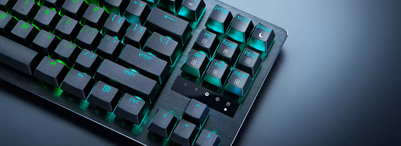 Gaming keyboard with black keys, blue and green backlit labels, and metallic finish on a dark surface.