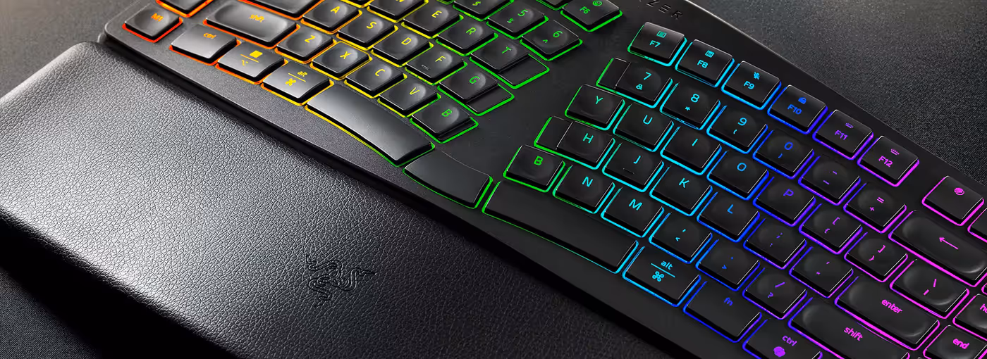 Close-up of a gaming keyboard with RGB backlit keys, positioned above a textured leather wrist rest embossed with the Razer logo.