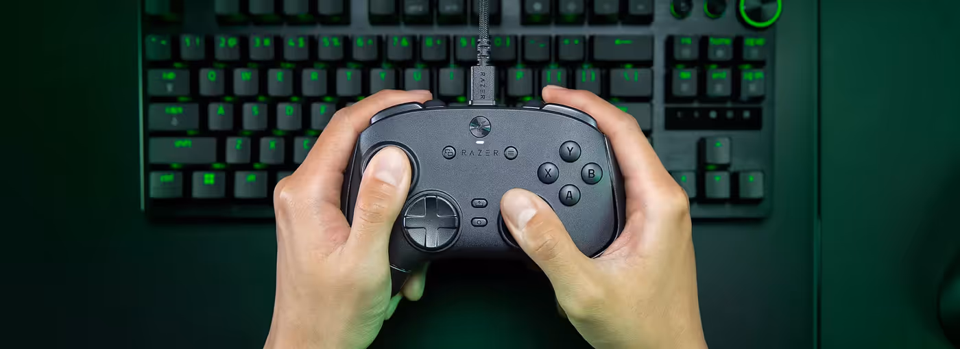 Hands holding a Razer gaming controller above a black gaming keyboard with green backlit keys.