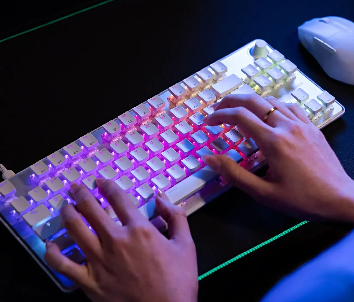A close-up of a backlit keyboard with colorful keys and a hand typing, alongside a computer mouse on a dark surface.